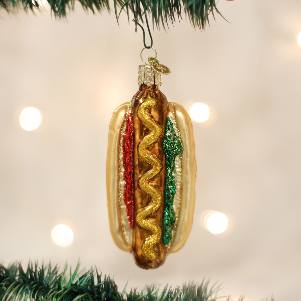 A hot dog-shaped Christmas ornament hanging from a green tree branch, with a gold glittery bun, brown sausage, red mustard, and green relish, set against a soft blurred background with warm bokeh lights.