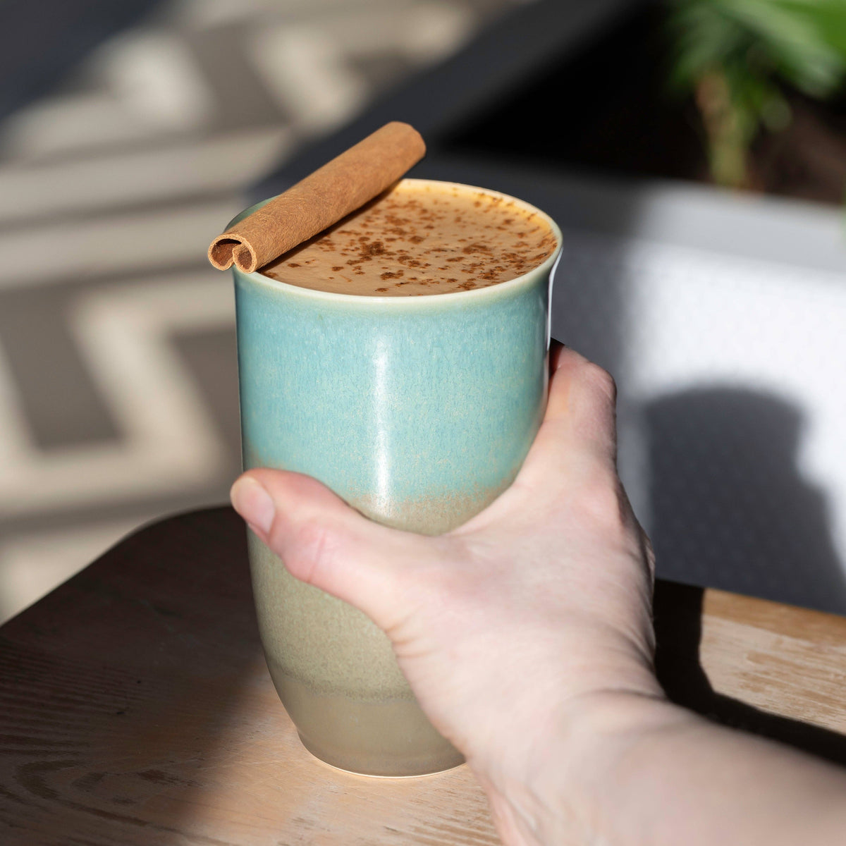 Hand holding a turquoise ceramic mug with a cinnamon stick on a wooden surface