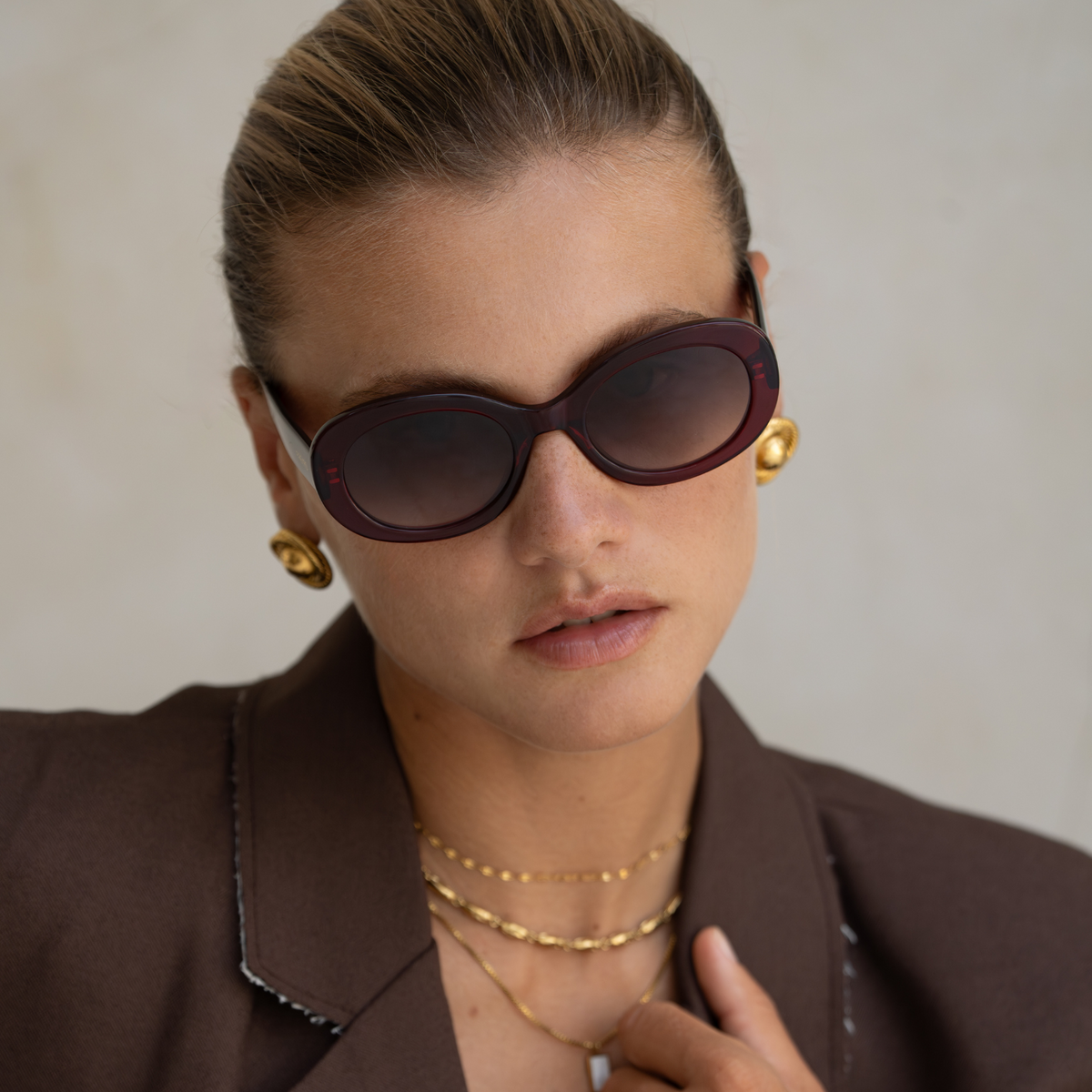 Woman wearing sunglasses, gold earrings, necklaces, and bracelets against a neutral background