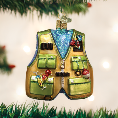 A Christmas ornament shaped like a gold fishing vest with green pockets, hanging from a tree branch against a softly blurred holiday background.