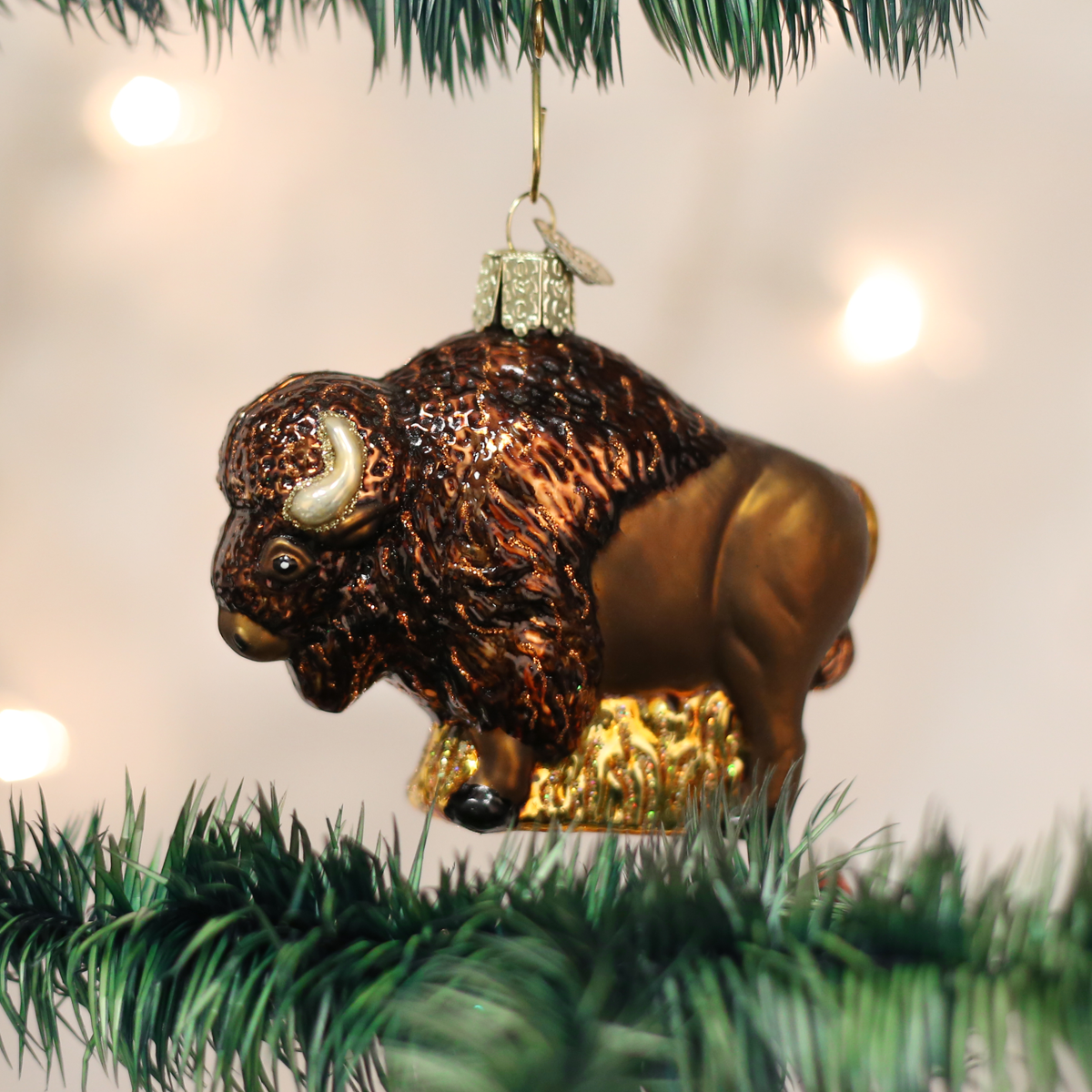 A metallic brown and gold bison Christmas ornament hanging from a green pine tree branch against a softly blurred white background with warm lights.