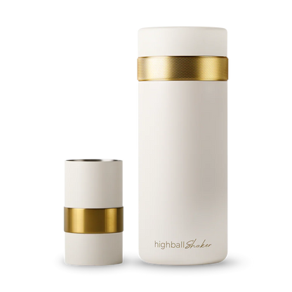 An off-white insulated bottle with a textured gold band and a matching off-white cap, shown on a clean white background. The shot glass is placed beside the shaker.
