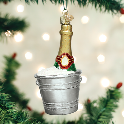 Decorative Christmas ornament of a champagne bottle in a bucket with lights and tree branches in the background.
