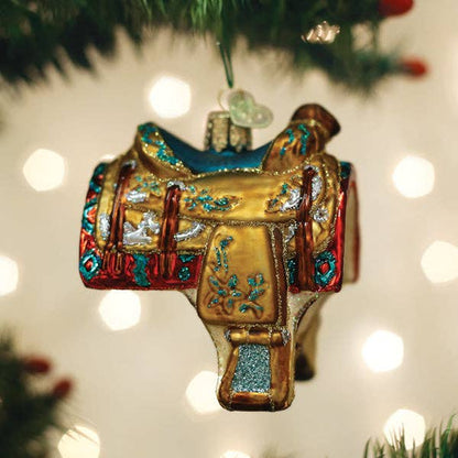 A decorative Christmas ornament shaped like a western saddle, hanging from a green tree branch with warm bokeh lights in the background.