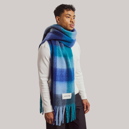 A man wearing a blue and teal plaid scarf with fringe, styled over a white long-sleeve shirt and dark trousers, photographed against a neutral light gray studio background.