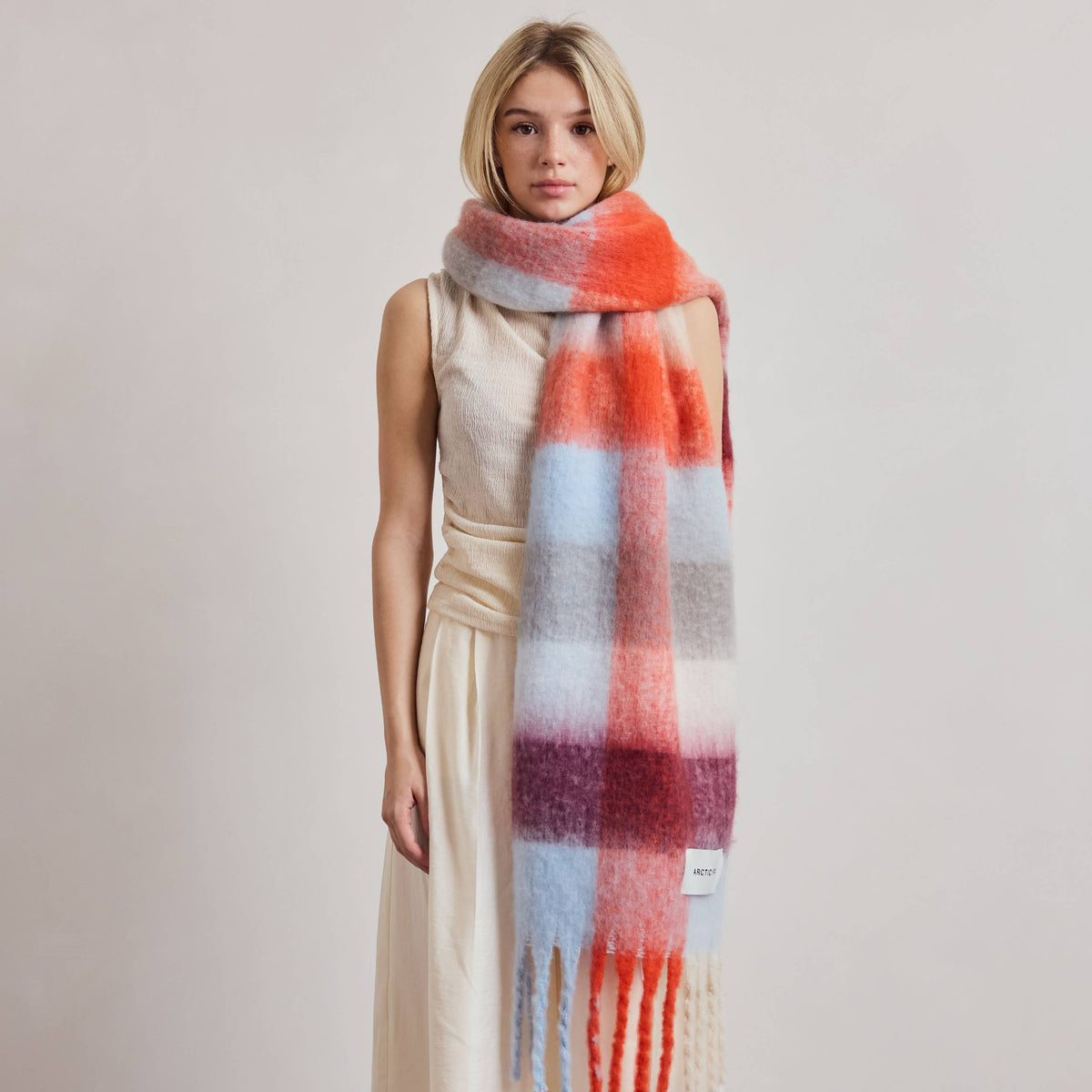 A model wearing a cream-colored sleeveless jumpsuit and a thick, fuzzy plaid scarf with orange, red, light blue, and purple stripes, standing against a neutral studio backdrop.