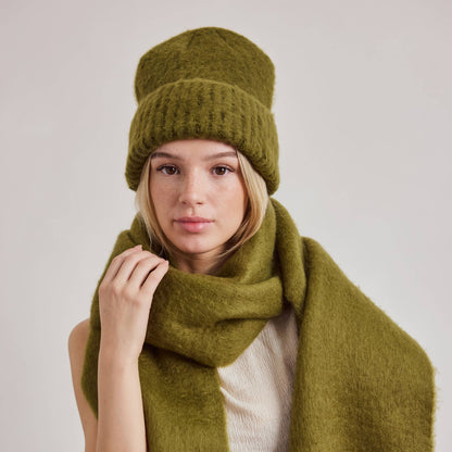 A woman wearing a matching olive green knit beanie, thick knit scarf, and cream ribbed top, posed against a light gray studio background. A small white brand tag on the scarf reads ANTON.