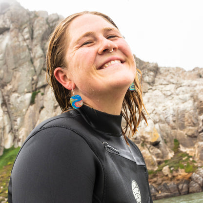 Person wearing a wetsuit and colorful resin earrings with a mountainous background