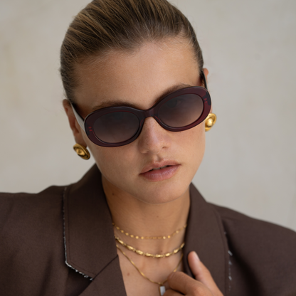 Woman wearing sunglasses, gold earrings, necklaces, and bracelets against a neutral background