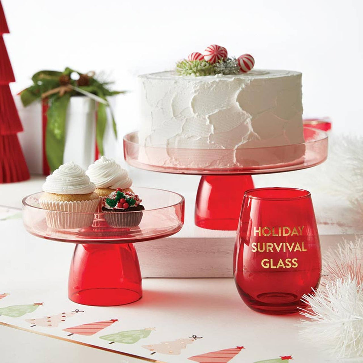 Red cake stand with a cake, cupcakes, and a 'Holiday Survival Glass' on a festive background.