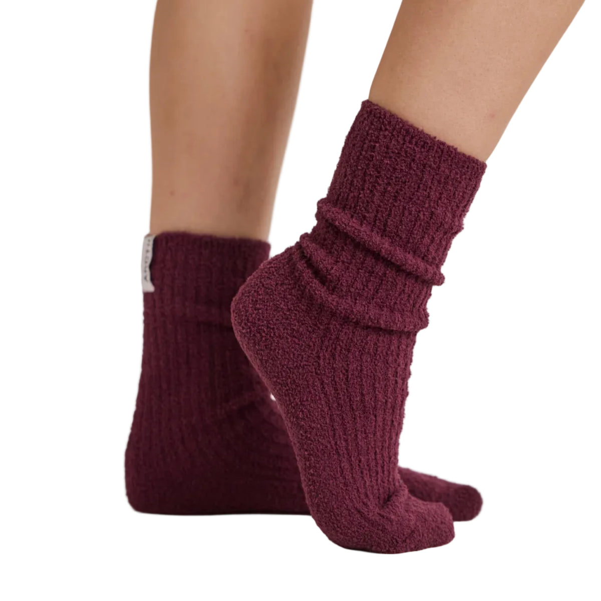 A close-up of a person's feet and lower legs wearing a pair of chunky, ribbed knit ankle socks in a deep burgundy or maroon color, set against a clean, light gray studio background.
