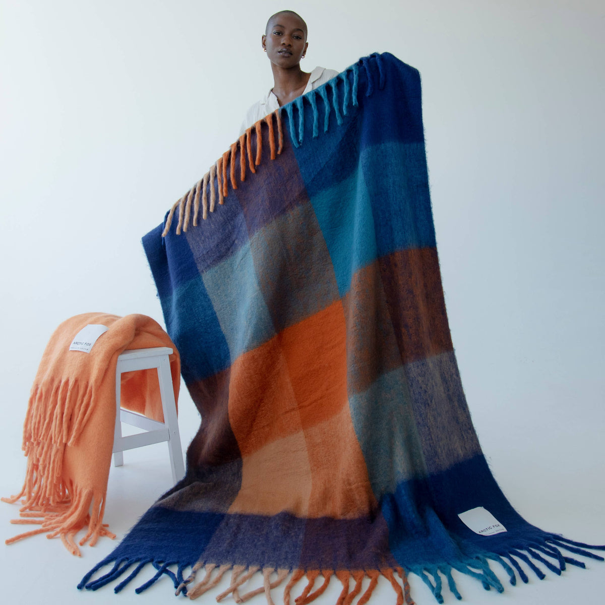 A person stands wrapped in a large woven throw blanket with fringe, holding it open to display its color-blocked pattern. An orange throw blanket rests on a small white stool to the left.