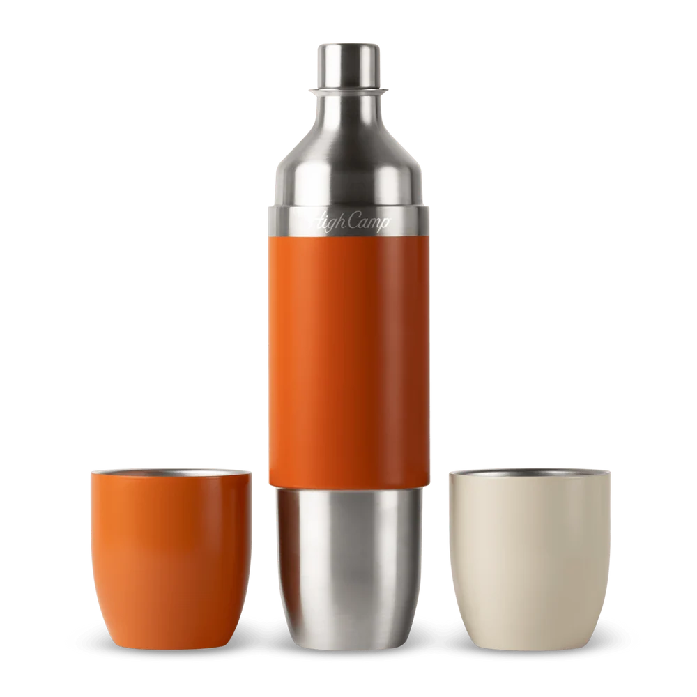 A stainless steel cocktail shaker with an orange sleeve, shown with two tumblers on a white background. One tumbler is orange and the other is a light cream color.